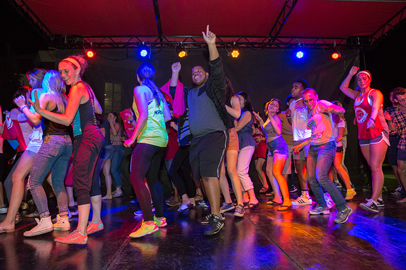 A large group of students dance on stage under colored lights